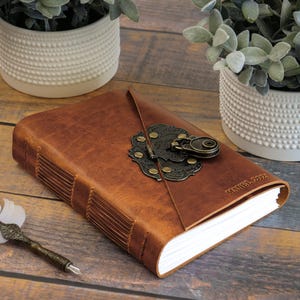 May include: A brown leather journal with a decorative metal clasp and the text "MANUEL 2022" embossed on the cover. A white feather quill pen rests beside the journal. The journal is surrounded by two white textured pots with green plants.