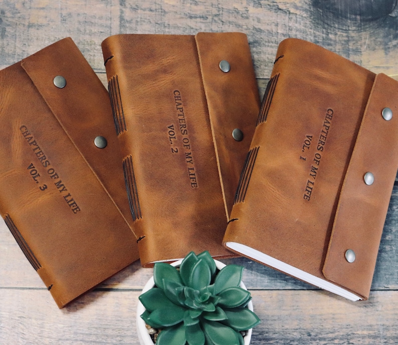 May include: Three brown leather journals with the text "Chapters of My Life" and volume numbers 1, 2, and 3. The journals are closed and have a rustic look.