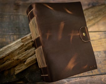 May include: A brown leather journal with a button closure. The journal is hand-stitched with beige thread and features a distressed finish. The spine is visible, showcasing the binding. The journal is resting on a piece of wood.