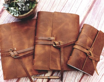 May include: Three brown leather journals with a rustic look. Each journal has a leather strap closure and a knotted tie. The journals are arranged in a size order from largest to smallest.