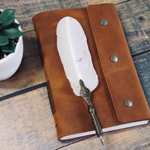 May include: A brown leather journal with a white feather quill pen. The journal has a flap closure with two metal poppers. The journal is lying on a wooden surface.