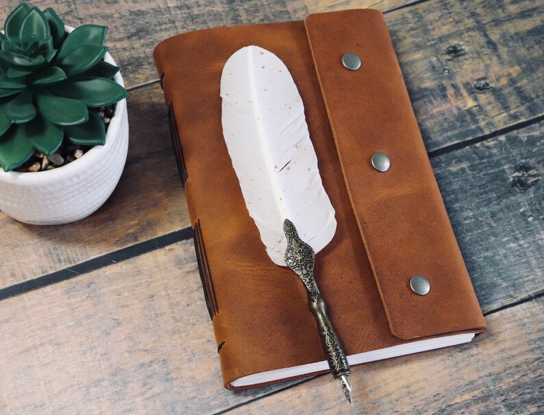 May include: A brown leather journal with a white feather quill pen. The journal has a flap closure with two metal poppers. The journal is lying on a wooden surface.