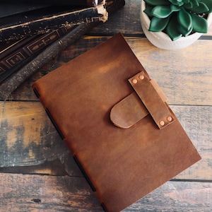 May include: A brown leather journal with a leather strap closure and copper rivets. The journal is lying on a wooden surface.