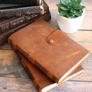 May include: Two brown leather journals with a button closure are stacked on a wooden surface. The journals have visible stitching along the spine. A stack of antique books and a small potted succulent are in the background.