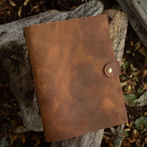 May include: A brown leather journal with a button clasp. The journal is rectangular and has a textured surface. The button clasp is gold-colored. The journal is sitting on a wooden surface.