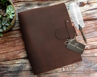 May include: A brown leather journal with a flap closure and a tag that reads "HANK & BELLE". A decorative feather quill pen rests beside the journal. The journal is on a wooden surface with a succulent plant.