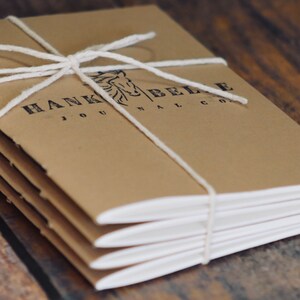 May include: A stack of brown kraft paper journals with white string tied around them. The cover has a black and white logo that says "Hank Belle Journal Co."