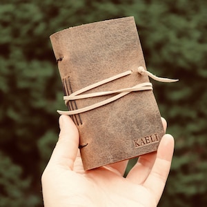 May include: A small, brown leather journal with the name "KAELI" embossed on the cover. The journal is bound with cream-colored string and has a rustic, worn appearance. The background is a blurred green.