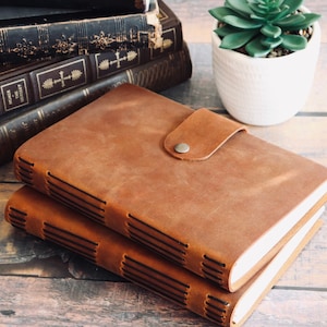 May include: Two brown leather journals with a stitched binding and a leather strap closure. The journals are stacked on top of each other on a wooden surface.