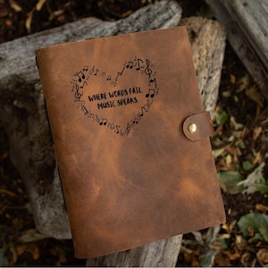 May include: A brown leather journal with a heart-shaped design made of musical notes and the text "WHERE WORDS FAIL, MUSIC SPEAKS." The journal has a button closure and is set against a natural background.