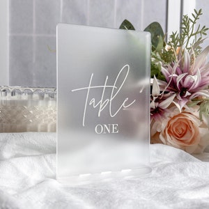 May include: A frosted acrylic table number sign with the word "table" and the number "one" written in white.