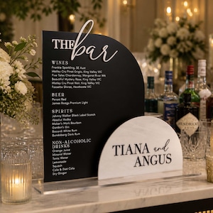 May include: A black bar menu sign with a semi-circular design, listing wines, beers, spirits, and non-alcoholic drinks. A smaller white sign displays the names "TIANA and ANGUS".