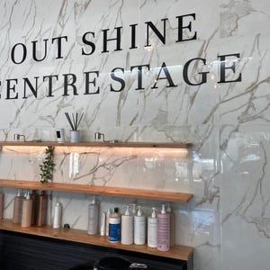 May include: Interior of a salon with a marble-effect wall displaying the text "OUT SHINE CENTRESTAGE". Wooden shelves hold hair care products and decorative items. The design is modern and clean, suggesting a professional beauty space.