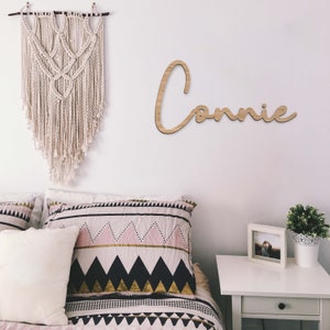 May include: A wooden sign with the name "Connie" written in a cursive font. The sign is hanging on a white wall above a bed with a white and pink patterned duvet cover.