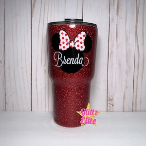 Może przedstawiać: Czerwony kubek z brokatem z czarno-białym wzorem głowy Myszki Minnie i imieniem Brenda. Kubek ma czarną pokrywkę.