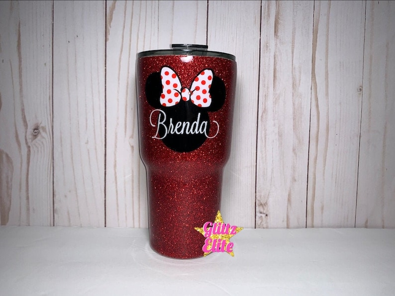 Może przedstawiać: Czerwony kubek z brokatem z czarno-białym wzorem głowy Myszki Minnie i imieniem Brenda. Kubek ma czarną pokrywkę.