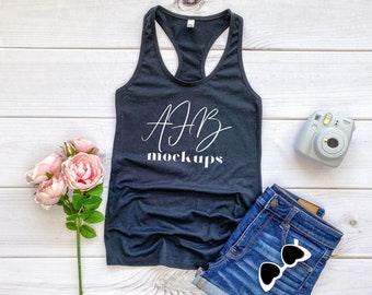 May include: A black racerback tank top with the text "AFB mockups" in white on a white wood background. The tank top is laying flat with a pair of blue denim shorts and a vintage camera beside it.