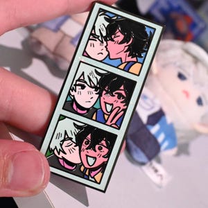 May include: A rectangular enamel pin featuring three comic-style panels. Each panel depicts two cartoon figures in various affectionate poses, with one figure having white hair and the other black hair. The pin has a light blue border.
