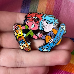 May include: Enamel pin featuring two cartoon characters hugging, each holding a skateboard. One character has red hair and the skateboard says "REKI". The other has blue hair and the skateboard says "LANGA".