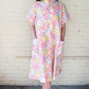 May include: A knee-length floral print dress in pink, blue, and yellow on a white background. The dress has short sleeves, a mandarin collar, and two front pockets. The model is wearing yellow sandals.
