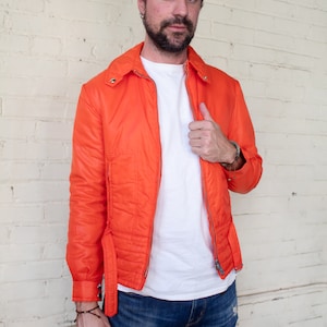 May include: An orange quilted jacket with a zip closure and a belt detail. The jacket is worn over a white t-shirt and paired with blue jeans. The person is standing in front of a white brick wall.
