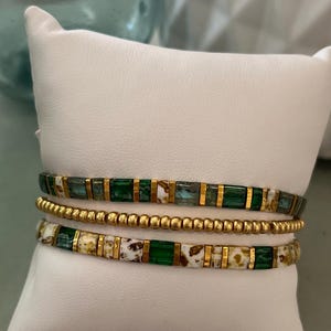 Op de afbeelding: Drie gestapelde armbanden met groene, gouden en witte kralen. De armbanden worden getoond op een wit kussen.