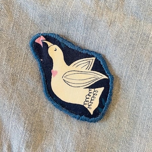 May include: A fabric patch featuring a white dove with black outline details, a pink heart, and a pink object in its beak. The dove is set against a dark blue background with a blue stitched border.