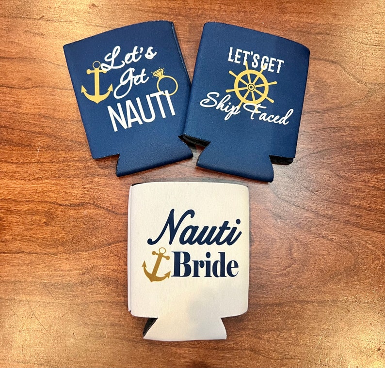 May include: Three navy blue and white can coolers with nautical-themed text and graphics. Two coolers read "Let's Get Nauti" and "Let's Get Ship Faced". The third cooler says "Nauti Bride" with an anchor graphic.