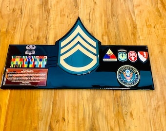 May include: A black and teal military plaque featuring a Sergeant Major rank insignia. The plaque includes military unit patches, ribbons, and the United States Army emblem. The plaque is displayed on a wooden surface.