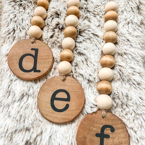 May include: Three wooden bead garlands with circular wooden pendants. Each pendant has a black letter: "d", "e", and "f". The beads are a mix of light and medium brown tones. The garlands hang against a fluffy, light-colored background.