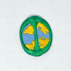 May include: An oval-shaped embroidered patch featuring a light blue background with yellow cloud-like shapes. A vertical green bar divides the center of the patch, and the entire design is bordered by a thick, stitched green edge.