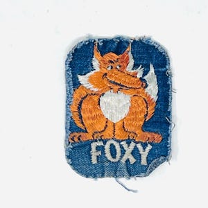 May include: A rectangular embroidered patch with a blue denim background featuring a cartoon fox with orange fur and a white chest. The fox is depicted in a seated position with a bushy tail. The word FOXY is stitched in white capital letters below.