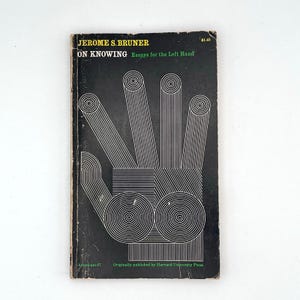 May include: A paperback book titled On Knowing: Essays for the Left Hand by Jerome S. Bruner. The black cover features a white line-art graphic of a hand composed of concentric circles and parallel lines, with the text printed in yellow and green.