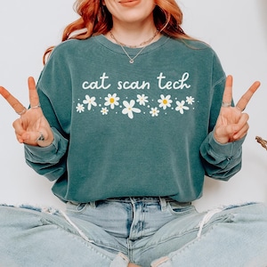 Peut inclure: Un sweat-shirt à col rond bleu sarcelle avec le texte blanc cat scan tech centré sur la poitrine, accompagné d'une rangée horizontale de fleurs marguerites blanches et jaunes imprimées juste en dessous du lettrage dans un design graphique.