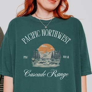Op de afbeelding: Een oversized tealgroen t-shirt met een witte grafische print die "Pacific Northwest Cascade Range" zegt met een zonsondergang en een berglandschap. De grafiek bevat ook de tekst "PNW" en "3000 MI".