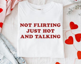 Könnte beinhalten: Ein weißes T-Shirt mit dem roten Aufdruck "NOT FLIRTING JUST HOT AND TALKING". Das Bild zeigt auch einen rosa Pullover mit roten Herzen, blaue Jeans und rote Herzdekorationen.