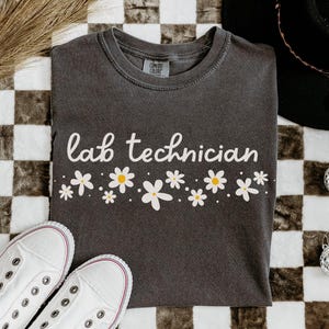 May include: Dark gray t-shirt with the words "lab technician" in white script, adorned with a row of white and yellow daisy graphics. White canvas sneakers and a black hat are also in the image. The shirt is made of a soft, comfortable material.