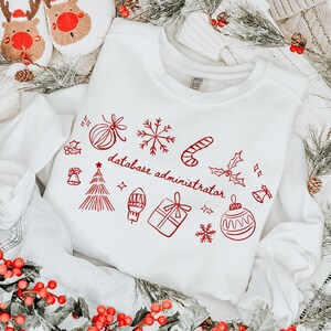 Könnte beinhalten: Ein weißes Langarm-Sweatshirt mit roten Weihnachtsgrafiken und dem Text "database administrator". Das Bild enthält festliche Dekorationen wie rote Beeren, Tannenzweige und Rentier-Hausschuhe.