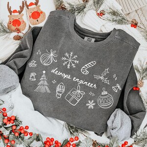 May include: A grey sweatshirt featuring white festive doodles and the words "devops engineer." The sweatshirt is styled with light-wash denim shorts, reindeer slippers, and a Christmas stocking. Red berry garland and pine branches add festive accents.