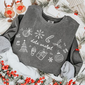 May include: A charcoal grey sweatshirt featuring the text "data analyst" and festive Christmas illustrations. The sweatshirt is styled with denim shorts, grey socks, and reindeer slippers. Decorative greenery and red berries are arranged around the items.