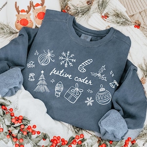 May include: A navy blue sweatshirt with white holiday-themed doodles and the text "festive coder." The sweatshirt is surrounded by Christmas decorations, including red berry garlands, a pair of reindeer slippers, and a white stocking.
