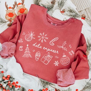 May include: A red sweatshirt with white holiday-themed graphics and the text "data engineer" is displayed with denim shorts, reindeer slippers, and a Christmas stocking. The scene is decorated with holly and pine branches.
