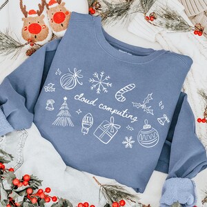 Op de afbeelding: Een blauwe sweatshirt met witte vakantie-thema doodles en de woorden "cloud computing". De afbeelding bevat kerstversieringen, waaronder rode bessen, dennentakken en rendierpantoffels.