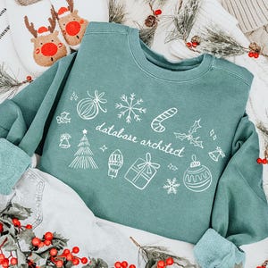 以下が含まれることがあります： 白いホリデーをテーマにしたグラフィックと「database architect」の文字が入ったティール色のスウェットシャツ。スウェットシャツはデニムショーツの上に置かれ、トナカイのスリッパとクリスマスの装飾が背景にあります。