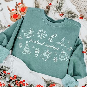 Può includere: Una felpa verde acqua con illustrazioni grafiche bianche e la scritta "frontend developer" è esposta su una superficie a righe bianche e nere. La felpa è circondata da decorazioni natalizie.