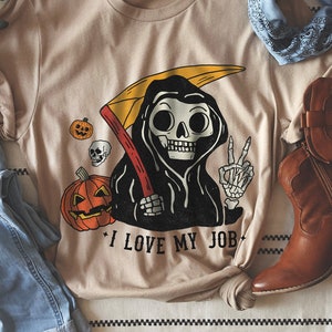 May include: A tan t-shirt with a graphic of a cartoon skeleton wearing a black robe and holding a scythe. The skeleton is holding up a peace sign and there are pumpkins and skulls around it. The text "I love my job" is printed below the graphic.