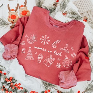 May include: A red sweatshirt with the text "women in tech" and holiday-themed graphics, paired with denim shorts and reindeer slippers. The image is decorated with red berries and pine branches, creating a festive holiday theme.