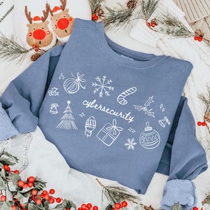 May include: A blue sweatshirt with the word "cybersecurity" and Christmas-themed doodles in white. The sweatshirt is laid on a white surface with a pair of reindeer slippers, denim shorts, and Christmas decorations.