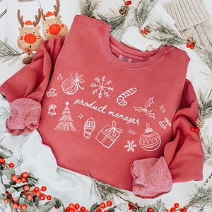 以下が含まれることがあります： 白いホリデーテーマのグラフィックと「product manager」の文字が入った赤いスウェットシャツ。スウェットシャツは、ライトウォッシュデニムショーツ、トナカイのスリッパ、クリスマスストッキングと組み合わせています。お祝いの緑と赤いベリーがホリデーシーンを完成させます。