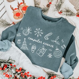 Könnte beinhalten: Ein teal-farbenes Sweatshirt mit dem Schriftzug "computer science" und weihnachtlichen Kritzeleien. Das Sweatshirt wird mit hellblauen Jeans, Rentier-Hausschuhen und einem Weihnachtsstrumpf kombiniert. Dekorative Girlanden mit roten Beeren und Tannennadeln runden die festliche Szene ab.